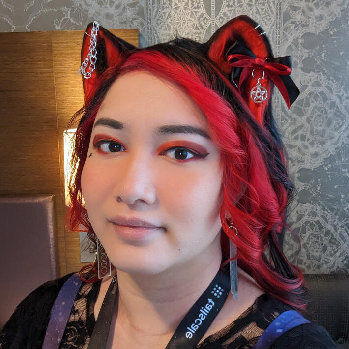 Headshot of a femme nonbinary person with red and black hair, smiling directly at the camera. They are wearing a black and red punk cat ears.
